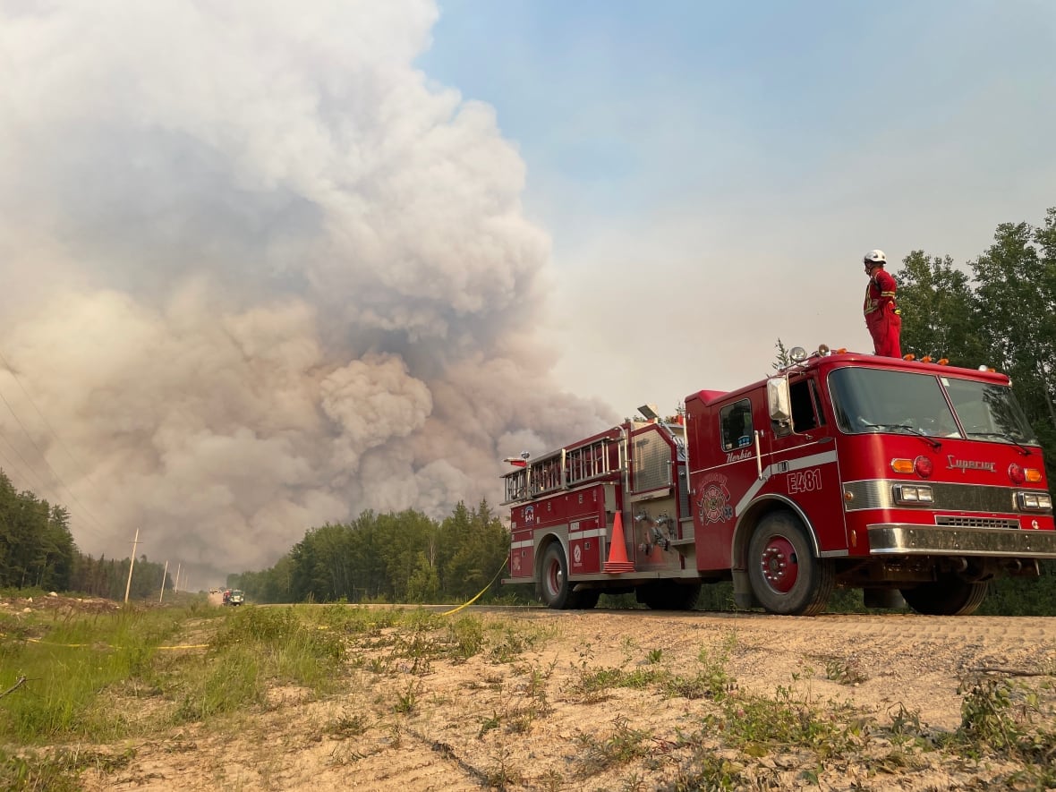 A firefighter stands on top of fire truck, looking out at large plume of smoke from a distant fire.