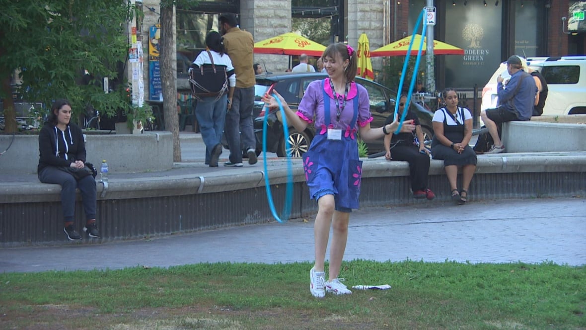 A person on blue and pink overalls spins blue hula hoops while performing on a grassy area near a stage.
