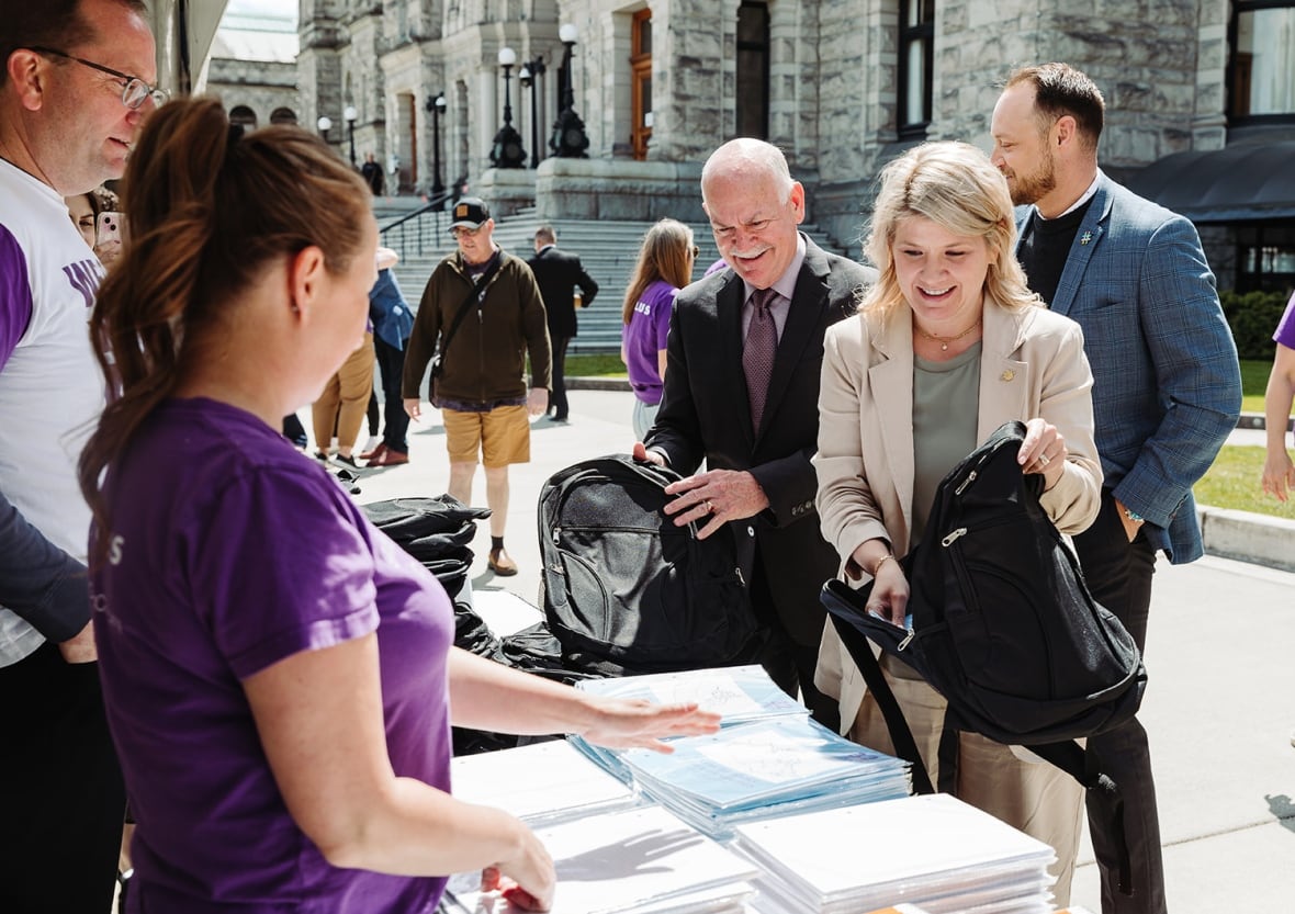 A woman flanked by people looks at a pile of reports outside the stone walls of the B.C. Legislature.