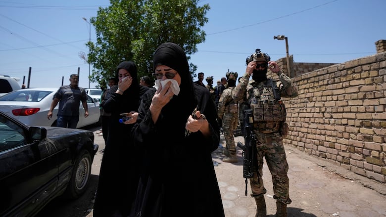 A woman in a religious headcovering wearing sunglasses holds a cloth to her face as men in camouflage uniforms are shown nearby.