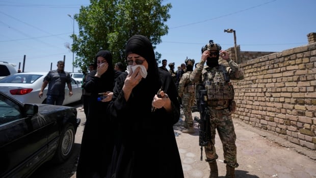 A woman in a religious headcovering wearing sunglasses holds a cloth to her face as men in camouflage uniforms are shown nearby.