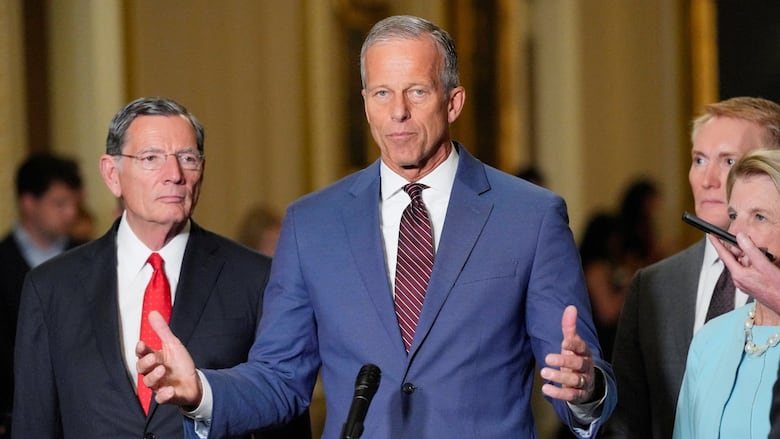 A cleanshaven shorthaired man in a suit and tie holds his hands apart while speaking at a podium while other people stand near him.