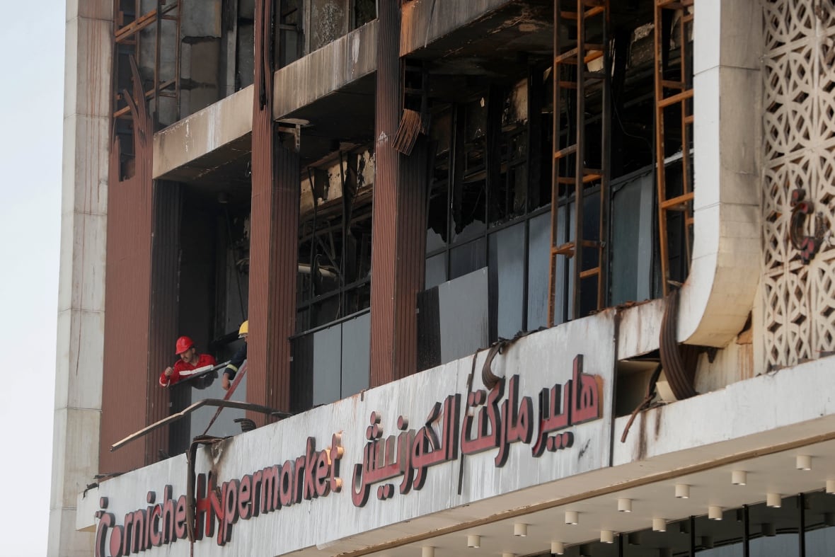 A closeup of damaged and blown-out windows of a building is shown, with a helmeted rescue worker seen through one of the windows.