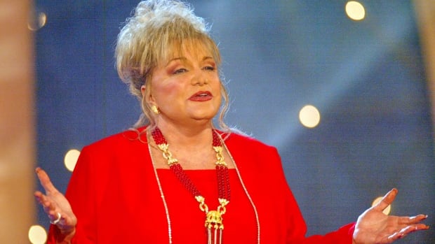 A woman with short blonde hair and a red jacket holds her hands up on stage.