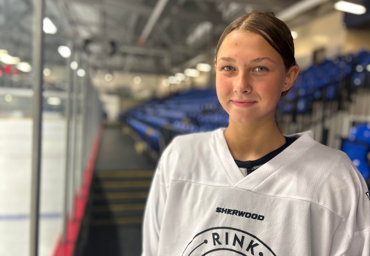 A teenaged girl dressed in a hockey uniform with her blonde hair pulled back in a ponytail smiles at the camera, with an ice rink behind her.