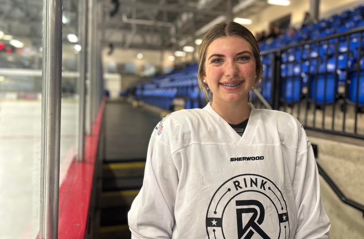 A teenaged girl with brown hair pulled back smiles at the camera, while wearing hockey uniform with an ice rink behind her.