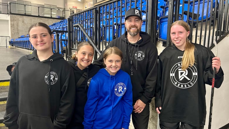 A man stands in the middle of a group of young girls dressed in hockey gear or sweaters with the Rink Rebels logo on it, and with bleachers in the background.