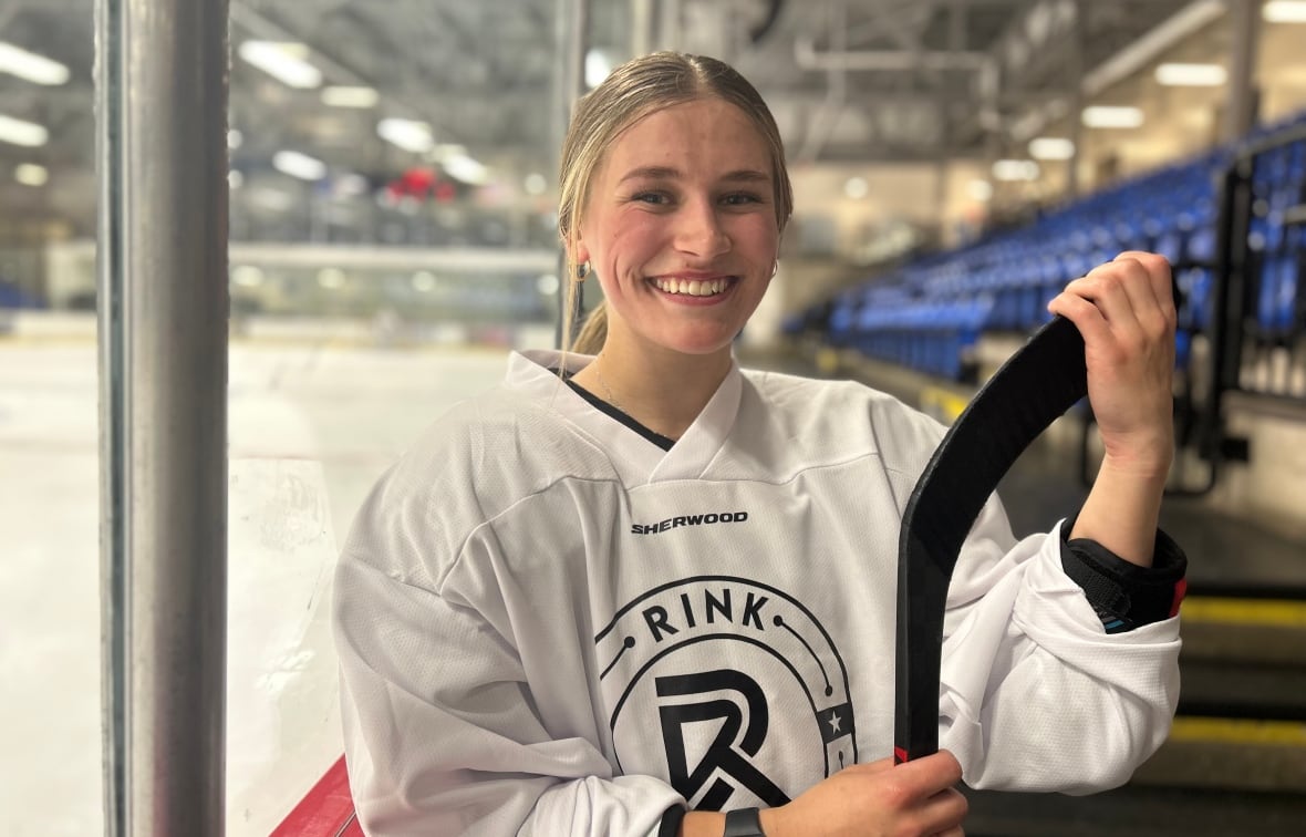 A young woman dressed in hockey gear smiles at the camera while holding a hockey stick with a rink behind her.