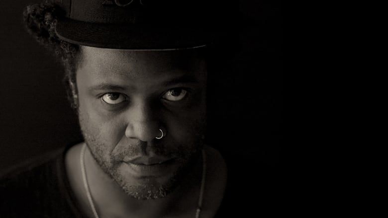 A Black man with a nose ring wears a hat and stares intently at the camera against a black background. 
