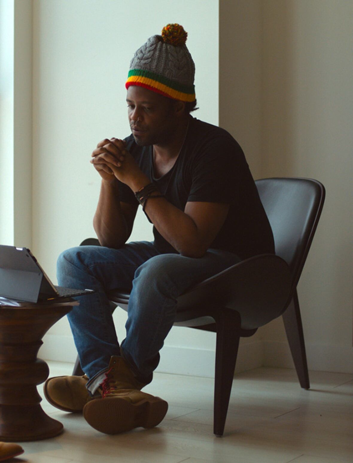 A Black man wearing a colourful wool hat sits in a chair watching a screen. 