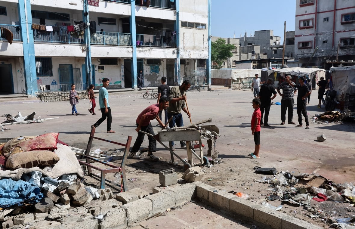 An urban square is shown, with men milling about and one using a wheelbarrow, as concrete debris on the ground is shown.