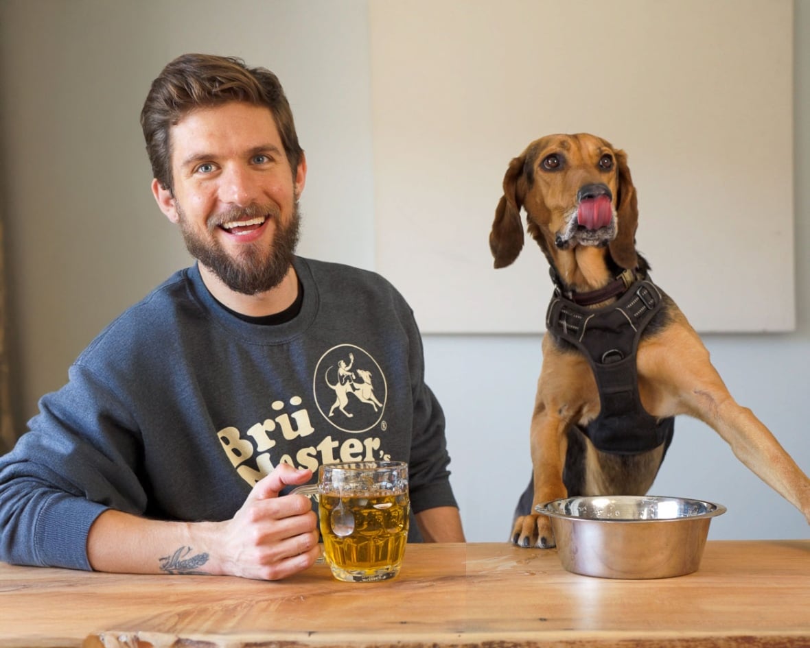 A man sits next to a dog. He is holding a "dog beer", the dog is licking his lips.