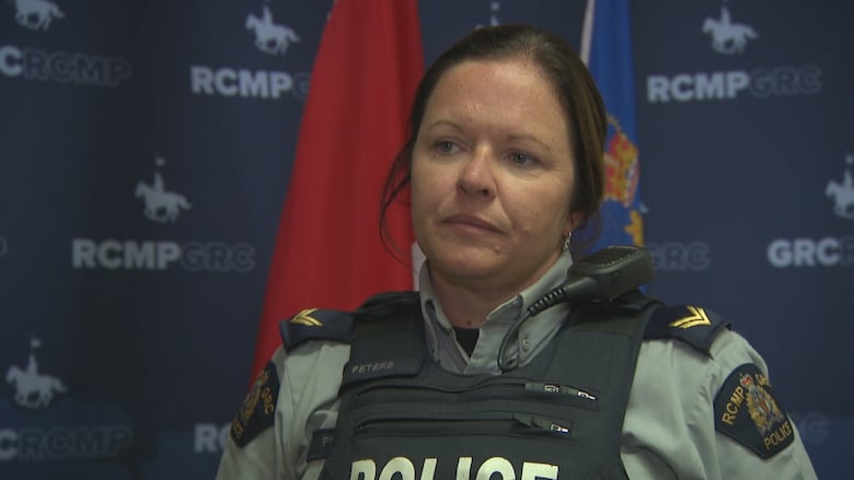 A police officer in an RCMP uniform stands indoors in front of RCMP-branded banners and two flags
