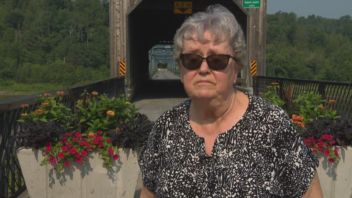 A woman with short grey hair wearing sunglasses, in front of a covered bridge with flower pots in front of it.