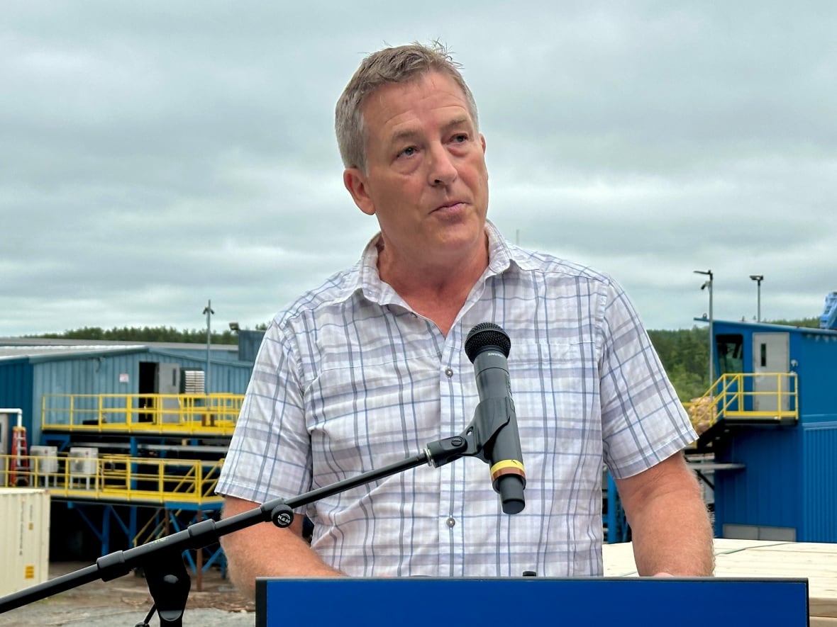 A man speaks into a microphone. A sawmill is in the background.
