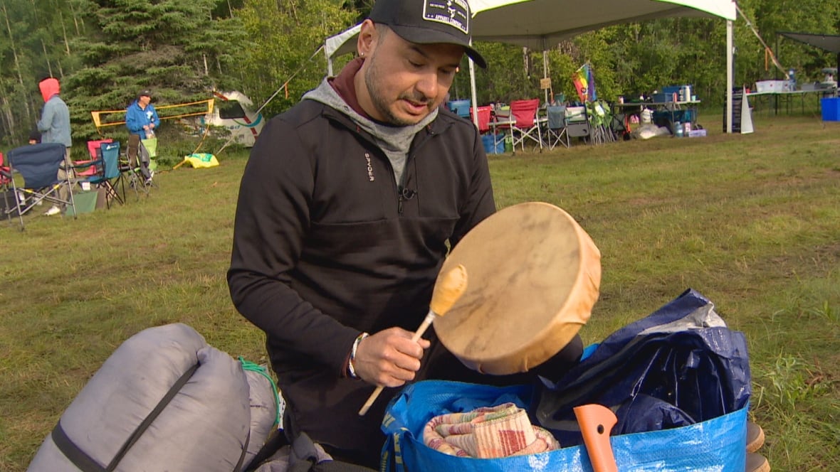 A person with a ball cap and black sweater hits a small hand drum.