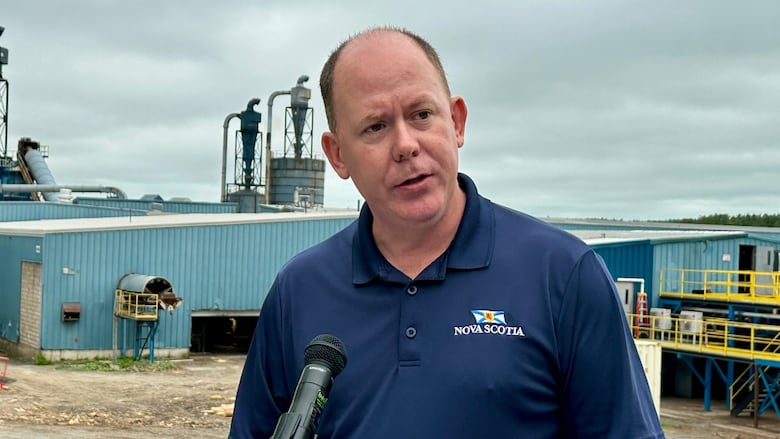 A man speaks into a microphone. A sawmill is in the background.