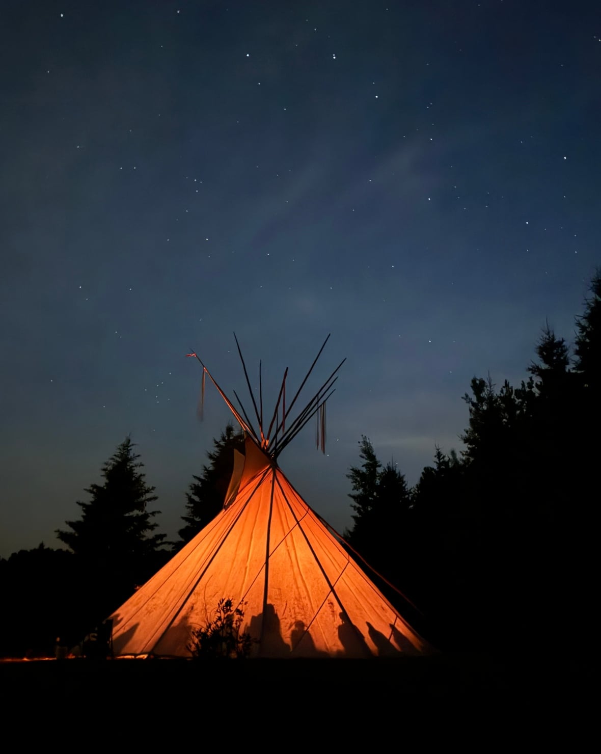 A teepee is lit up at night. Shadows of people inside are visible.