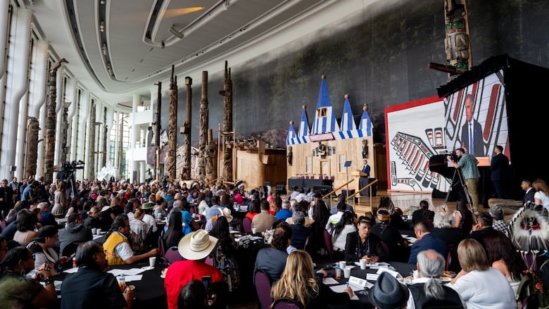 A wide shot of a full room with chiefs sitting at tables looking as Mark Carney speaks on  a stage. 