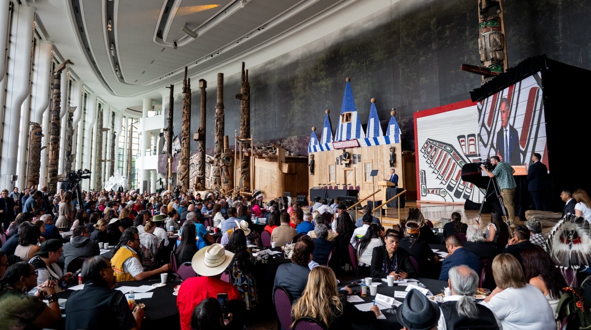 A wide shot of a full room with chiefs sitting at tables looking as Mark Carney speaks on  a stage. 