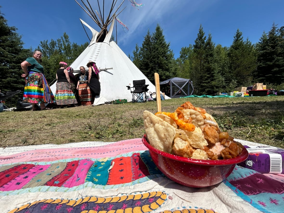 A bowl full of food is on a blanket. In the background a teepee and people can be seen.