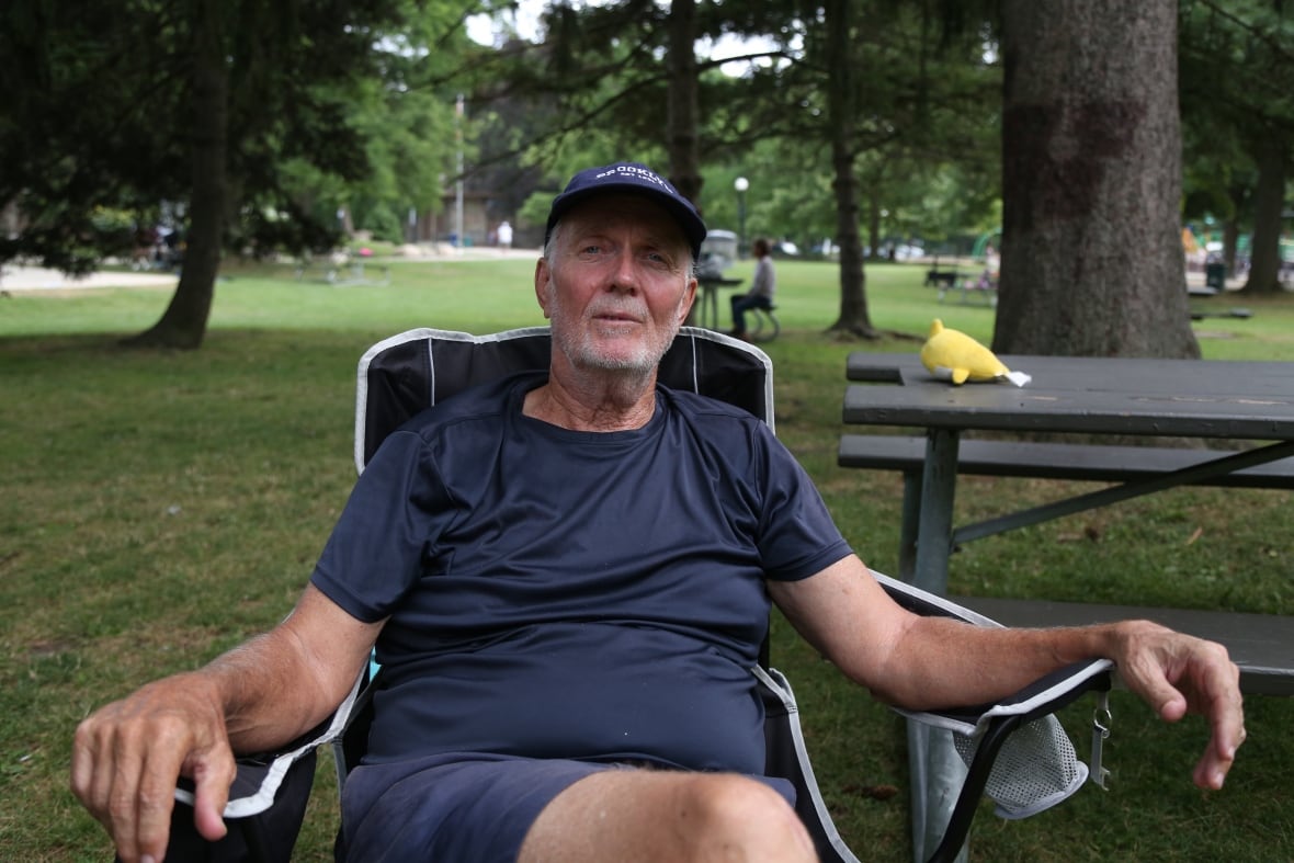 Person wearing a cap, seated in a lawn chair, looking at the camera, with both arms on respective arm rests.