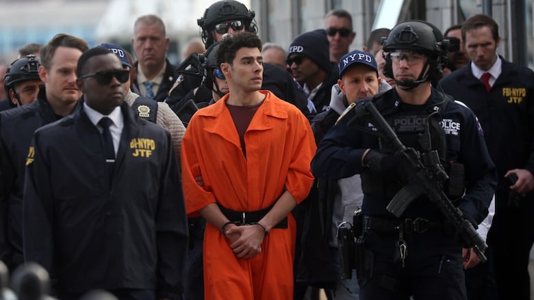 A man in an orange jumpsuit is flanked by a crowd of police, one of whom has a huge gun.
