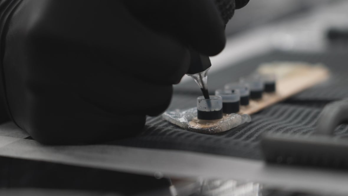 Close up image of a line of small ink pots, with a gloved hand dipping a tattoo needle in one of them