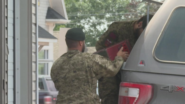 Two men in green camouflaged military uniforms remove equipment from the back of a grey truck.