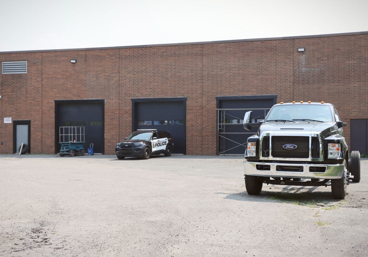 A black and white police car is parked in front of a garage door entrance to a large red brick building with a large black tow truck also parked out front.