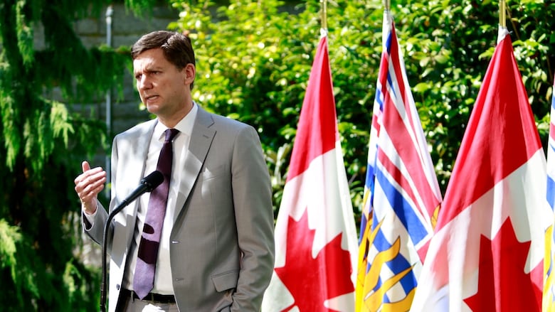 A man in a grey suit stands at a podium in front of Canada and B.C. flags.