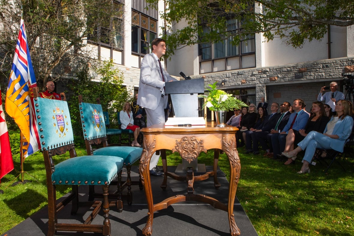 A man stands at a podium in front of a group of people. To his right is a table and two blue velvet chairs.
