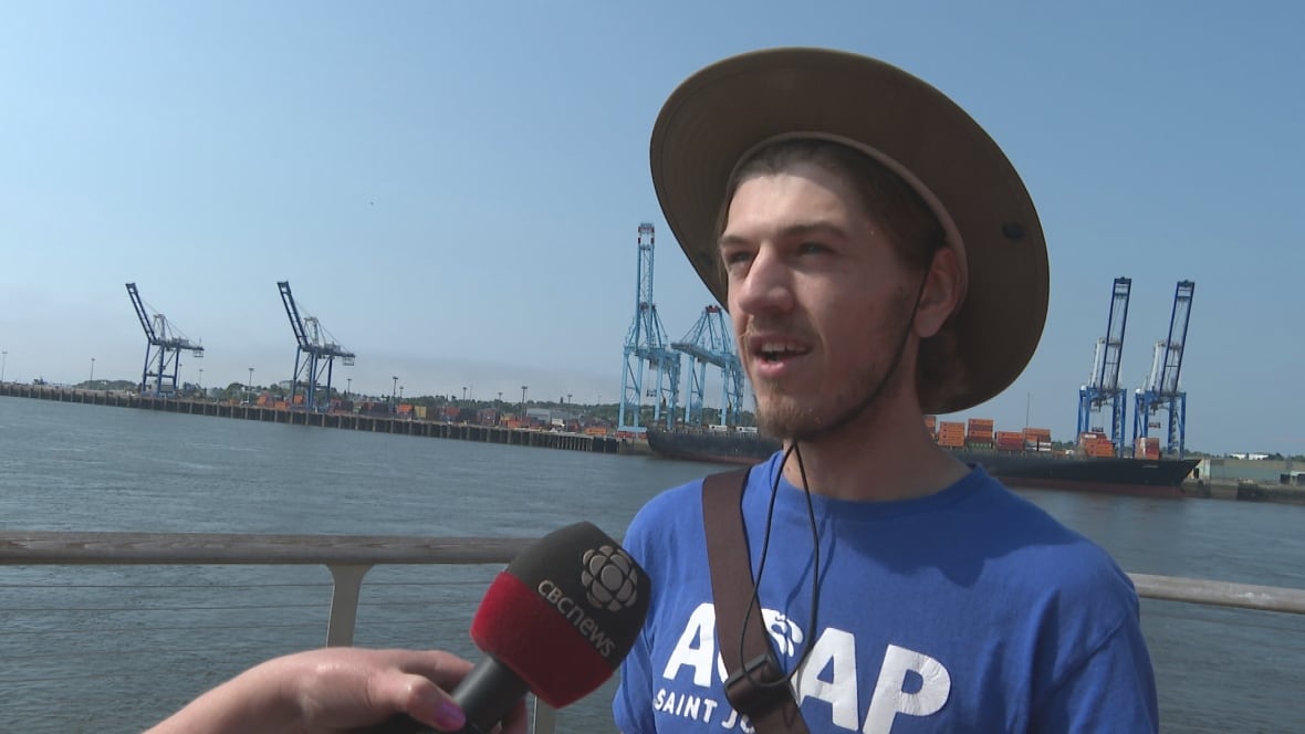 A man in a hat standing in front of cranes overlooking an ocean harbour.