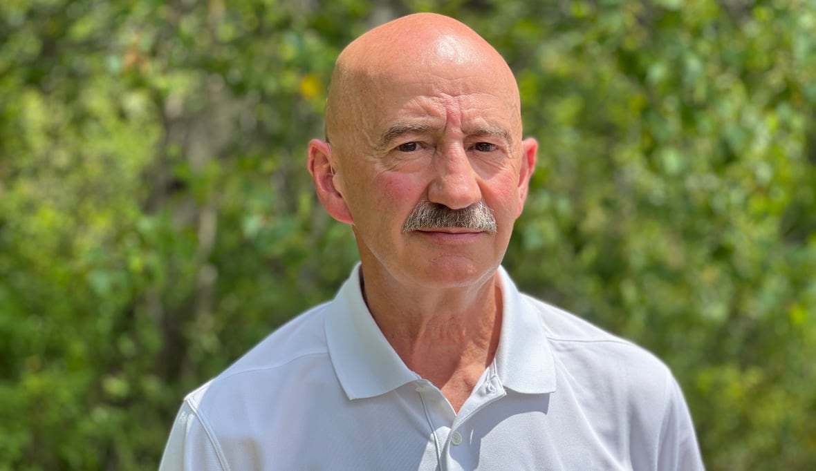 A white bald man in a white polo shirt with HALIFAX in blue on the chest stands outside, with green trees in the background