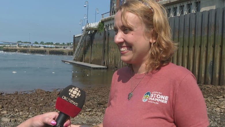 A smiling woman stands of the shore of an ocean harbour.