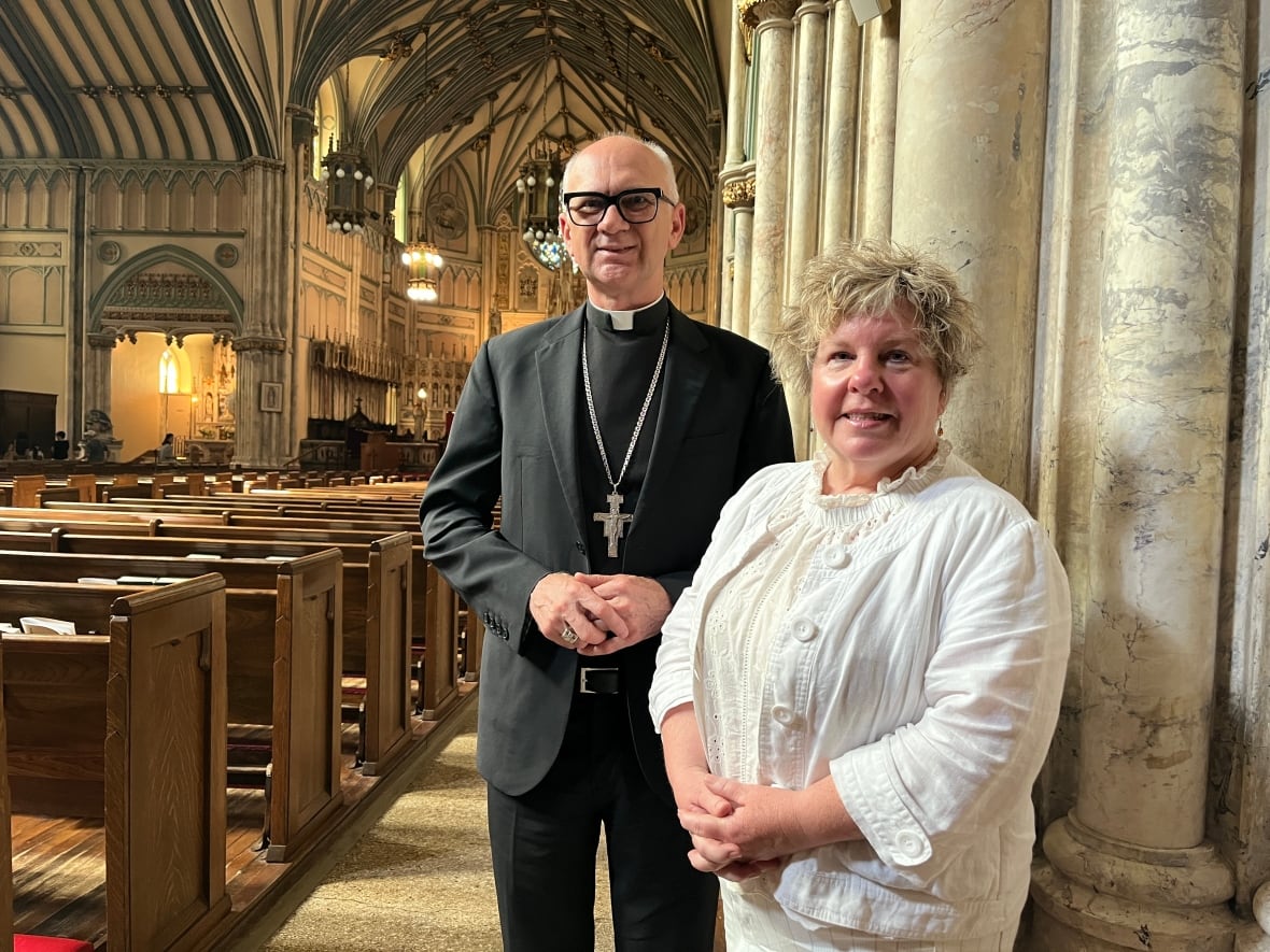 A man in a black suit and white clerical collar stands next to a woman with short curly hair in an ornate church. 