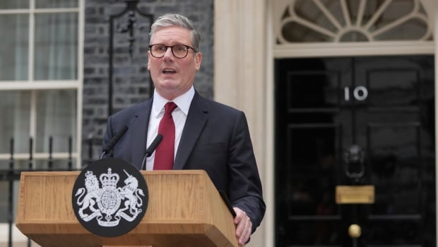 A man in a suit and red tie speaks into a microphone at a podium in front of a house.