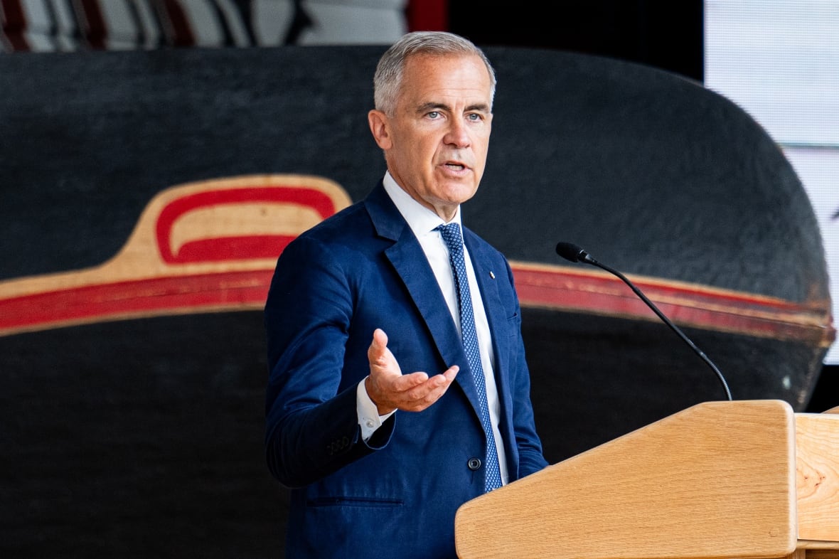 A man with short grey hair wearing a blue suit and blue tie gestures with his right hand while speaking at a podium.