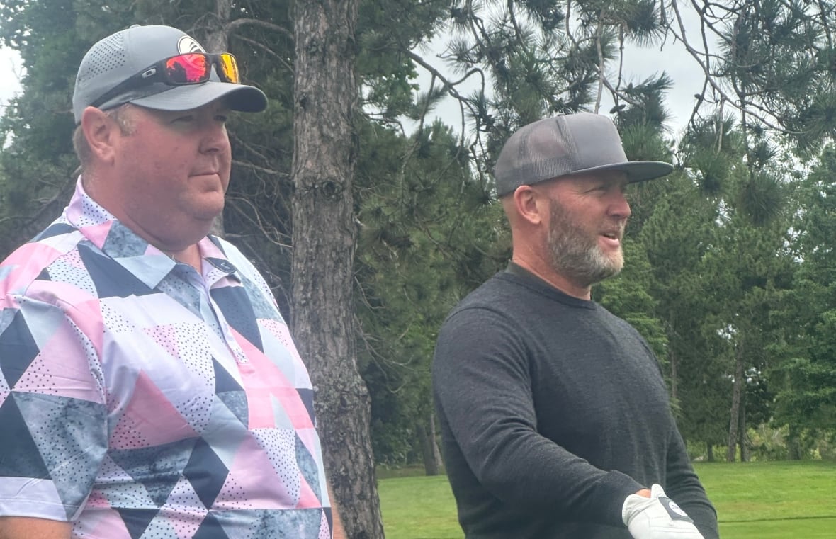 Two men in golf attire standing side by side with their golf clubs waiting to play golf.