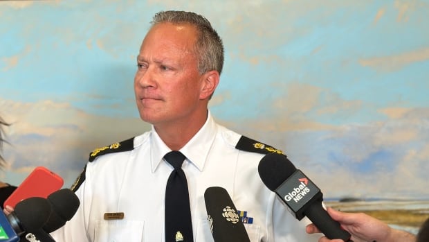 A police officer wearing a white button-up shirt and a tie stands in the middle of people holding microphones.