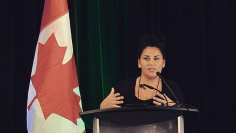 A woman speaks at a podium while other panellists and speakers listen. Next to her is the red and white flag with a maple leaf. 