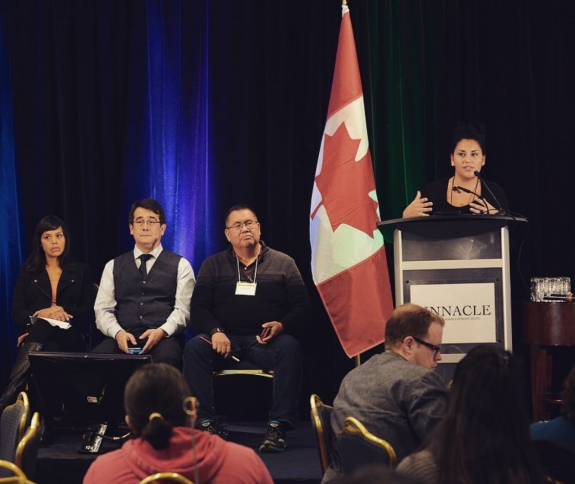 A woman speaks at a podium while other panellists and speakers listen. Next to her is the red and white flag with a maple leaf. 