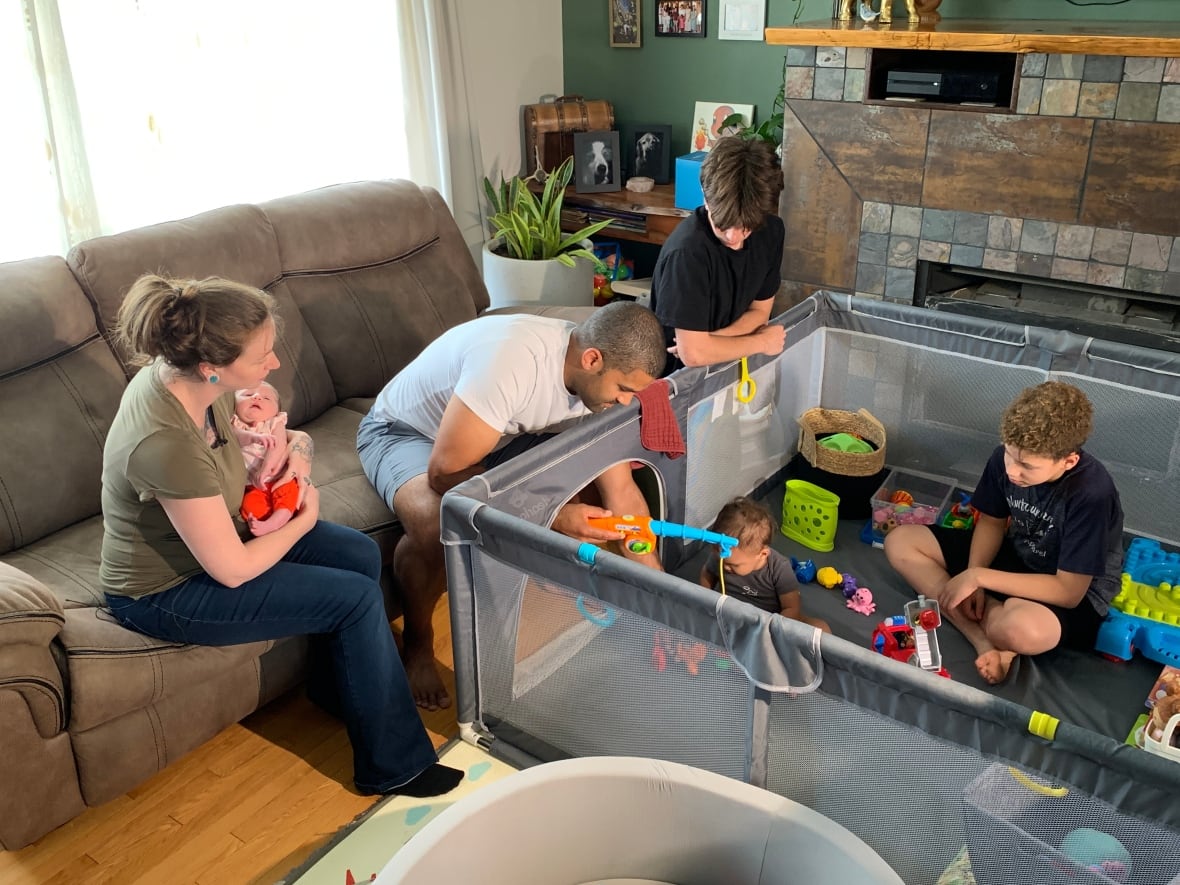 A woman holds a baby while a man sits on a couch and three other children play in the room.