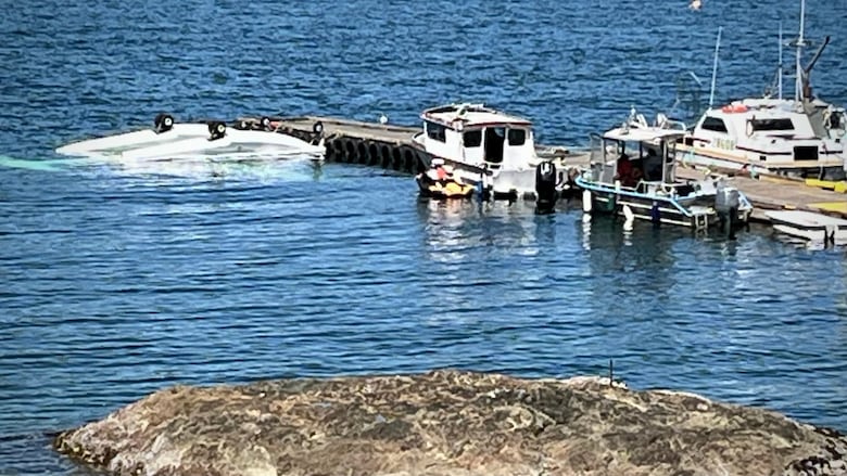 Several vessels moored on a dock next to an overturned plane whose wheeled pontoons are sticking out of the water.