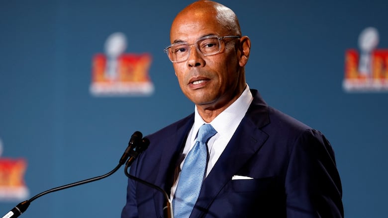 A man wearing a suit and glasses is shown standing behind a microphone.
