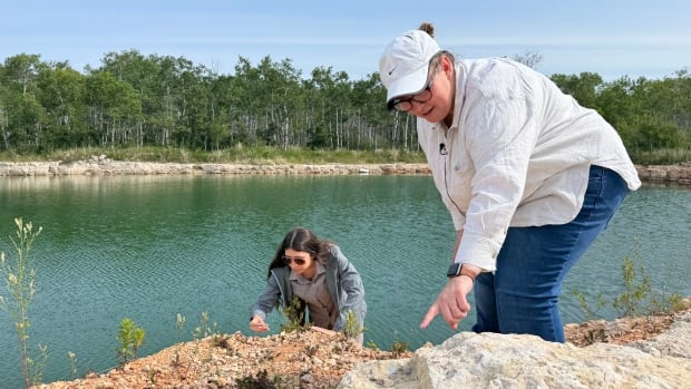 Two women stare intently at a rocky hill next to a body of water, with the woman on the right pointing at the rock.