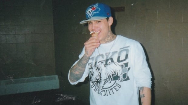 A man with tattoos and a Toronto Blue Jays cap eats an ice cream indoors.