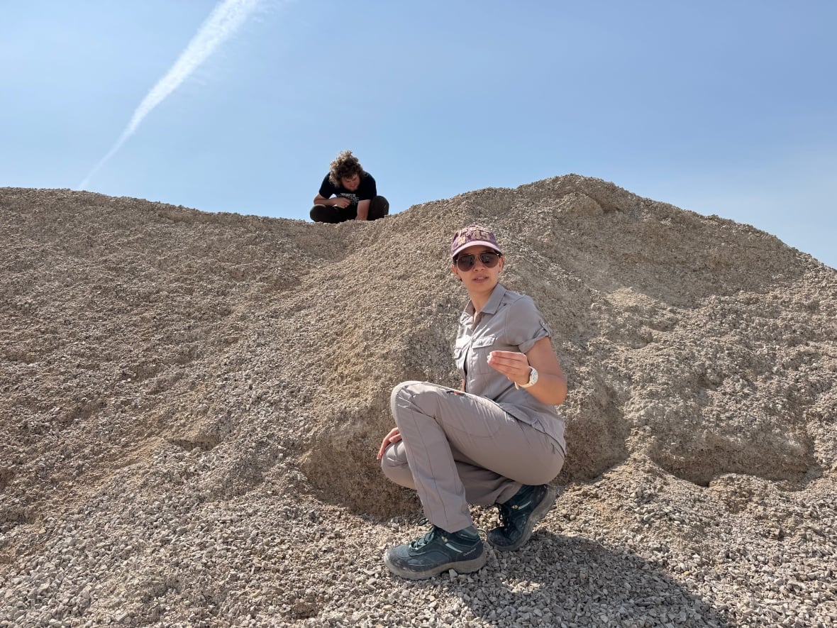 Une femme accroupie sur une colline recouverte de gravier tend une pierre vers la caméra, tandis qu'un homme accroupi au sommet de la colline rocheuse regarde le sol.