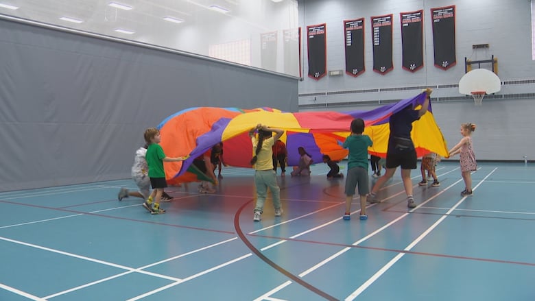 Children in a gym play under a multi-coloured piece of fabric.