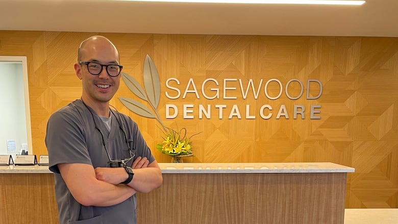 Dr. Brian Yim, a dentist, stands with a friendly expression in the reception area of Sagewood Dental Care. He is wearing professional scrubs, and the modern, clean design of the office is visible behind him.